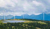 Wind turbines and solar panel installations in front of mountain slopes under a clear sky.
