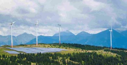 Wind turbines and solar panel installations in front of mountain slopes under a clear sky.