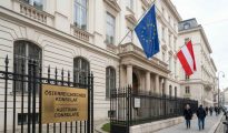 A real photograph showing the grand, classic white stone architecture of an Austrian Consulate building behind a black iron fence. A prominent gold sign reads "ÖSTERREICHISCHES KONSULAT / AUSTRIAN CONSULATE,"