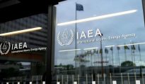 Glass entrance doors displaying the International Atomic Energy Agency (IAEA) emblem and text, with national flags visible in the background reflection at the Vienna International Centre.